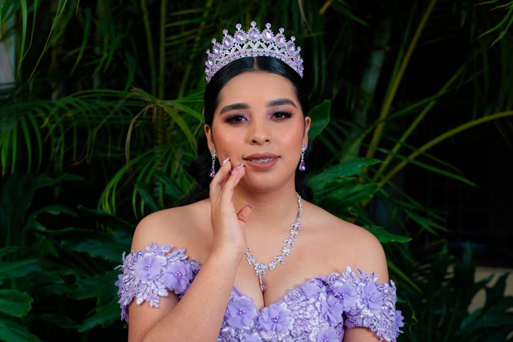 A beautiful woman wearing a lavender gown and tiara, standing gracefully with lush greenery in the background.